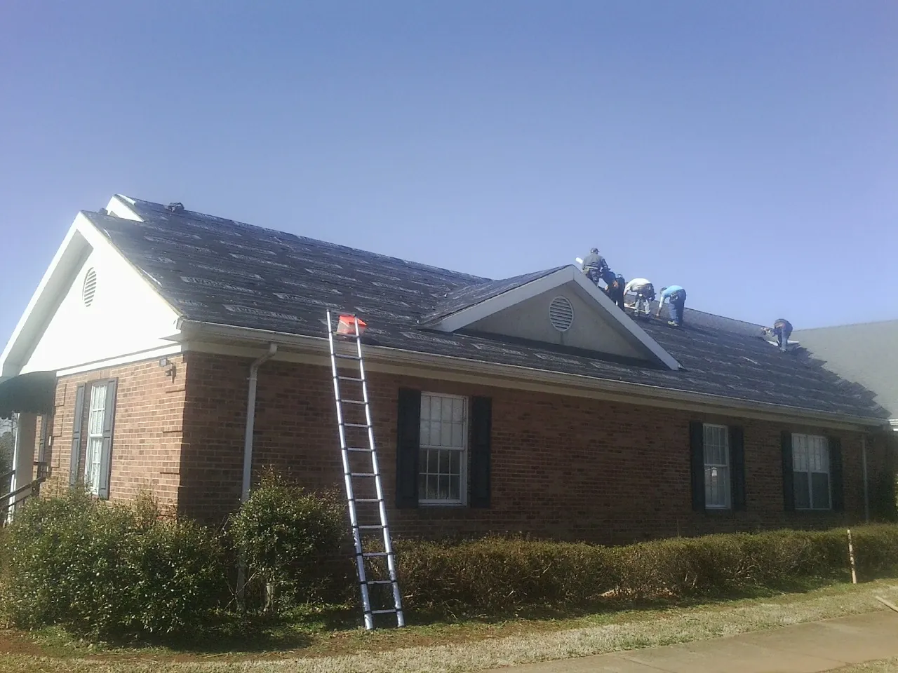 Crew tearing shingles off a brick ranch roof with a ladder set — Metal Roof Repair work by our crew in Mount Upton