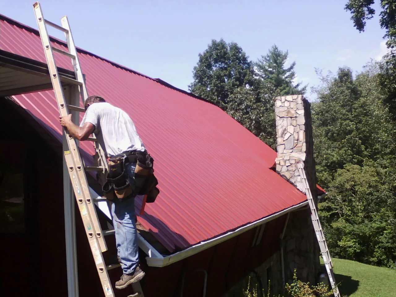 Roofer on a ladder beside a red metal roof and stone chimney — Soffit & Fascia Repair by Roofing Brothers in Mount Upton